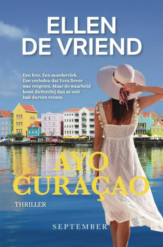 Een vrouw in een witte kanten jurk en brede hoed staat bij kleurrijke gebouwen aan zee in ayo curaçao