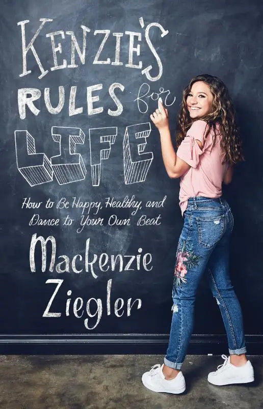 A woman in a pink ruffled top and floral embroidered jeans stands by a chalkboard of kenzies rules for life and dance
