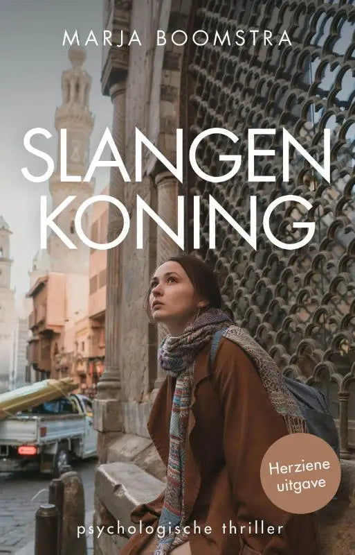 Een vrouw met donkere haren draagt een bruine jas en kleurrijke sjaal voor een sierlijke stenen poort in slangenkoning