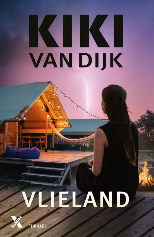 Het boekcover van Vlieland door Kiki van Dijk toont een vrouw in zwart op houten terras voor verlichte a-frame cabin bij dramatische lucht